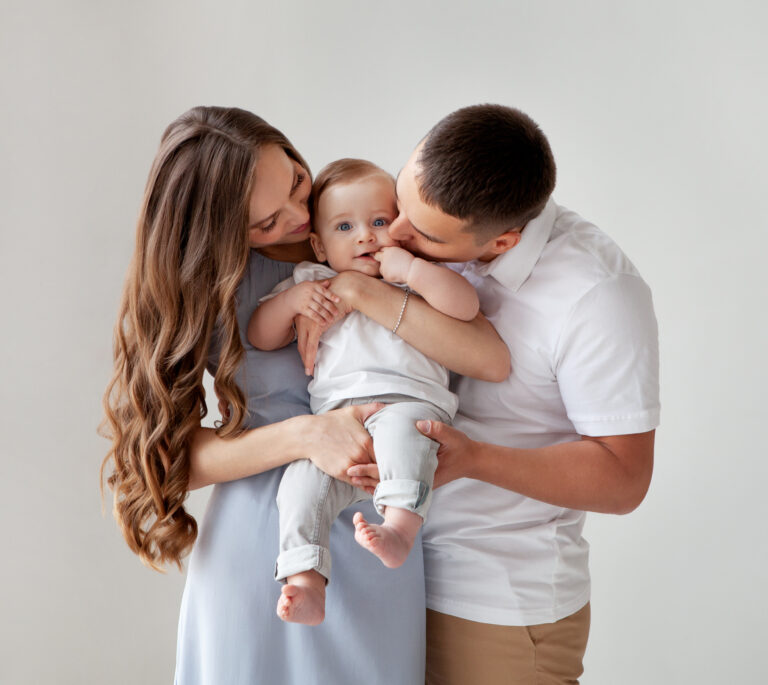 A woman and man stand together, holding and cuddling a baby who is looking at the camera. All three wear light-colored clothing against a plain background.