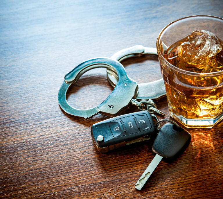 A glass of whiskey with ice, a pair of handcuffs, and a car key fob set on a wooden surface—reminding you to consult an OVI lawyer Dayton if you’re facing DUI charges.