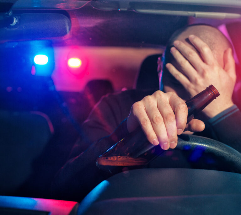 A person in a car holds a beer bottle and covers their face with one hand as police lights flash outside, capturing the tense moment of a Dayton drunk driving arrest.