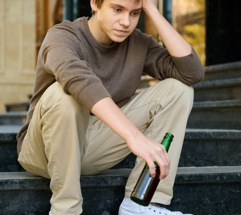 A young person sits on outdoor steps, holding a beer bottle, looking down with a thoughtful or worried expression—perhaps reflecting on the risks of underage drinking arrest in Dayton. Dressed in light pants, a brown sweater, and white sneakers.