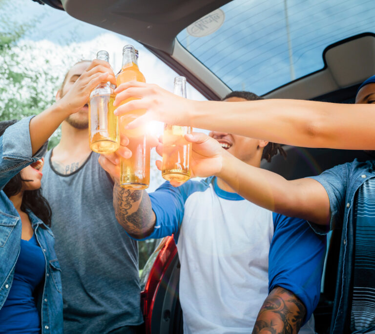 Four people clinking beer bottles together outdoors near an open car trunk, smiling and enjoying a sunny day—remember, OH underage OVI laws strictly prohibit anyone under 21 from drinking alcohol.